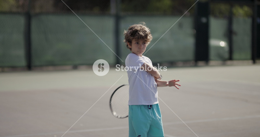 An enthusiastic young child plays tennis on an outdoor court, concentrating deeply as he prepares to hit a ball.