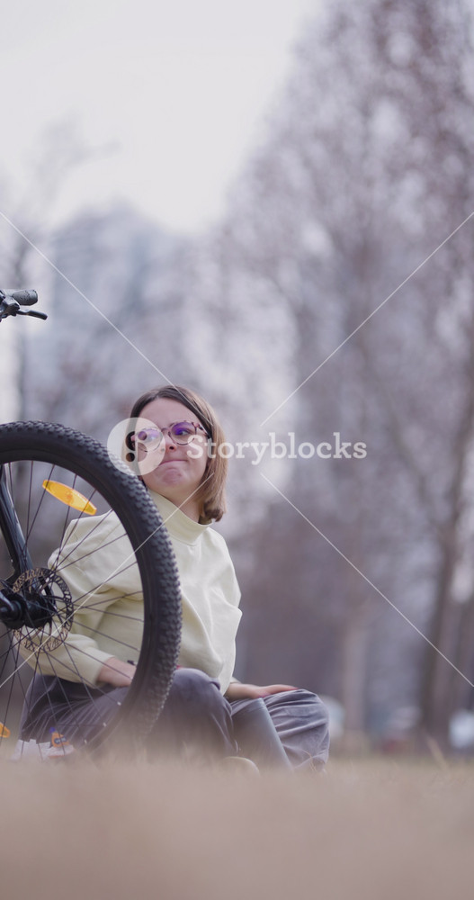 Relaxed young woman sits cross-legged in a park, talking on mobile phone beside her orange mountain bike, displaying a moment of leisure in