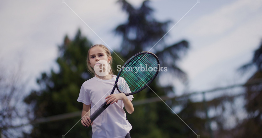 A focused young girl in a white t-shirt plays tennis, swinging her racket at a ball on a sunny outdoor court.