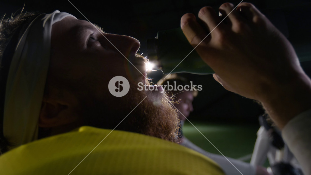 Side close-up of face of young bearded Caucasian male American football athlete pouring water from bottle in mouth and drinking during rest break, illuminated by spotlight with lens flare