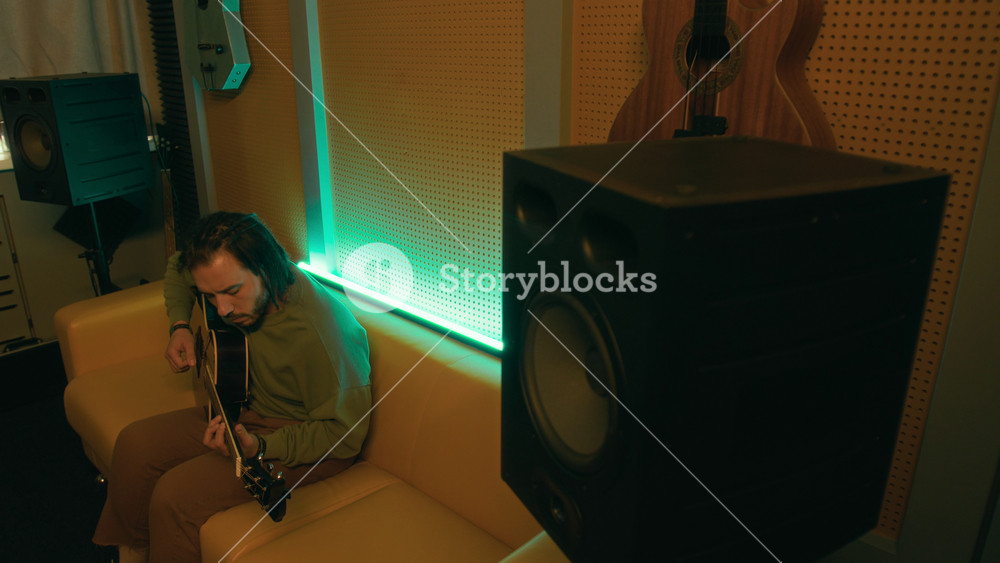 High angle side shot of young adult Caucasian man sitting on leather couch, playing and tuning acoustic guitar, turning tuning pegs on headstock in dim neon lit studio