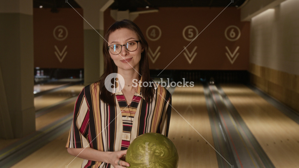 Portrait of Caucasian young zoomer woman with glasses holding bowling ball and smiling at camera in bowling alley. Perfect for promoting indoor sports, active lifestyle, and weekend leisure