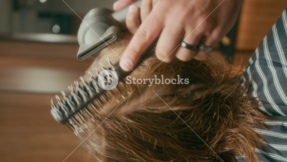 Vertical close-up of hands of anonymous barber styling male clients red curly hair with hairdryer and brush to achieve extra volume and bounce during haircut appointment