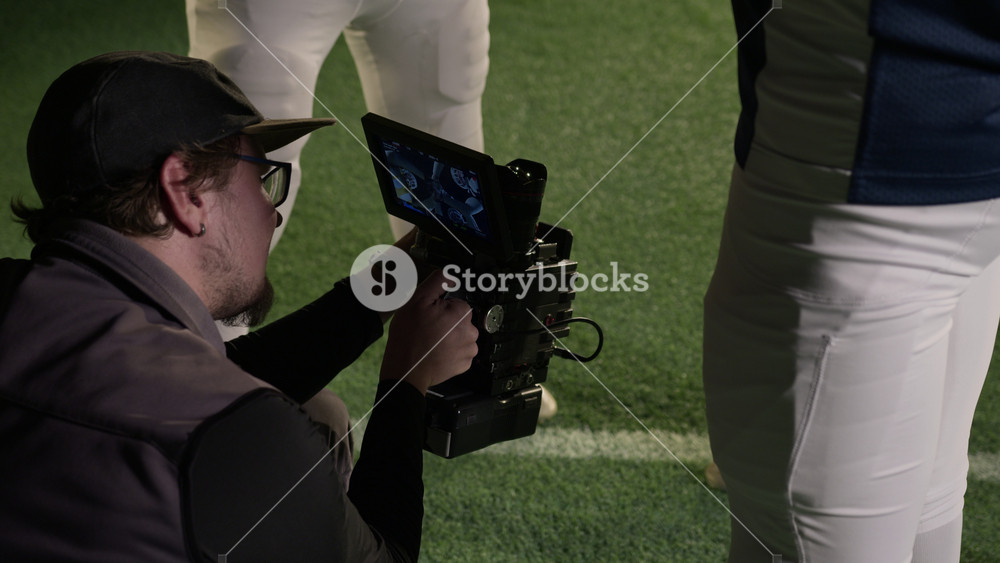 Medium shot of skilled professional Caucasian male videographer crouching on ground at sports stadium, filming gridiron football players hugging before game from ground up, focus on shot in viewfinder