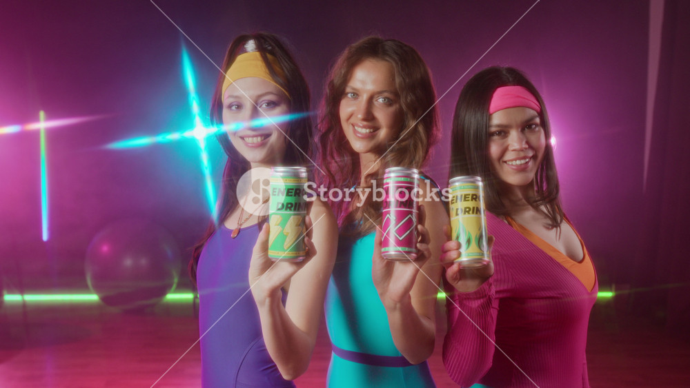 Medium portrait of three happy athletic diverse girls, wearing vintage bright leotards, headbands holding cans of energy drinks, looking at camera, after aerobics workout in gym studio