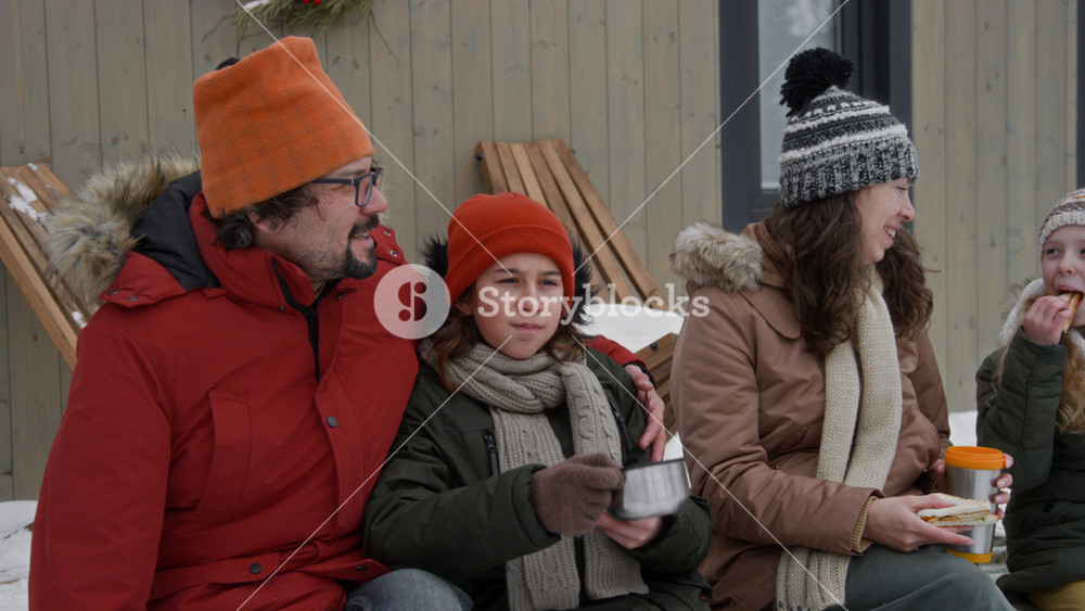 Medium shot of loving Caucasian father with limb difference disability, in warm jacket and hat, sitting on porch of holiday cabin, hugging teenage son, while mother and daughter snacking on sandwiches