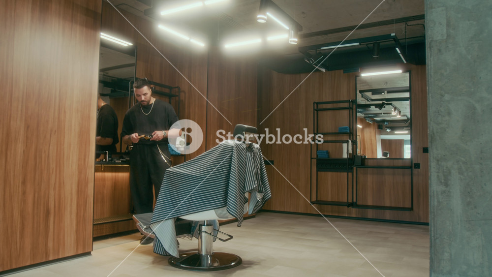 Wide shot of young Caucasian barber with tattoos standing in barbershop, cleaning tools, mature Asian male customer walking in, greeting with handshake, sitting in chair and discussing haircut request