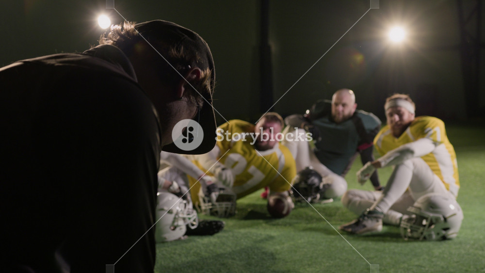 Medium rear behind scenes shot of videographer shooting portrait of American football amateur club team resting on green turf at stadium, for promotion or television program