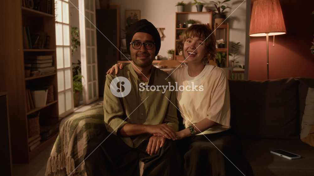 Medium portrait of diverse young adult couple of Caucasian blonde lady in white t-shirt and Indian man in turban and glasses holding hands on sofa, smiling and looking at each other and at camera