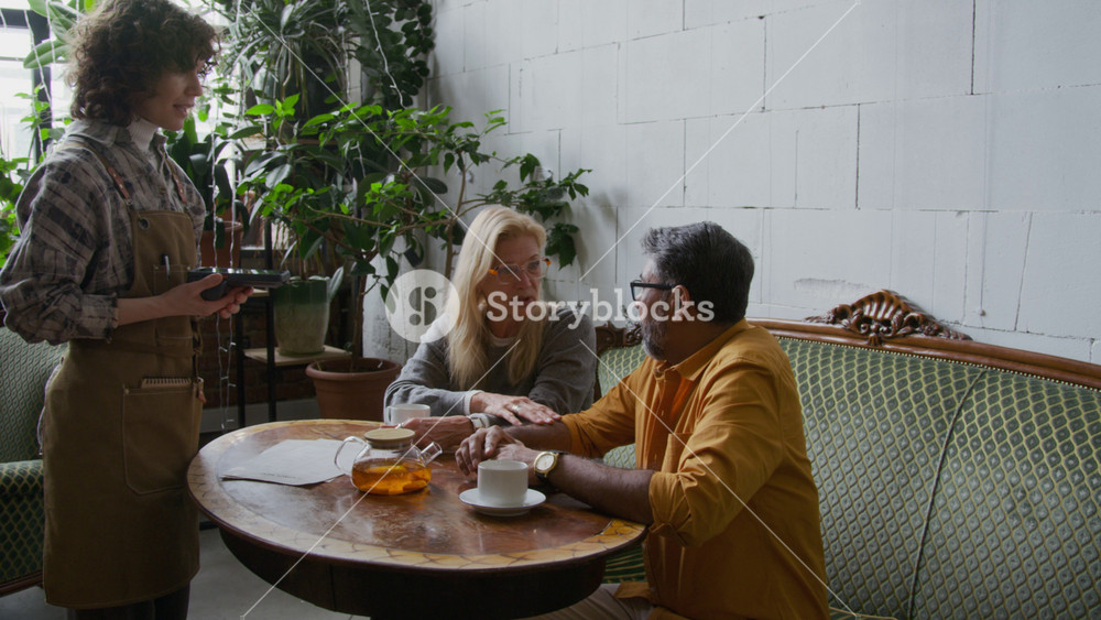 Medium full shot of happy multiethnic couple, mature Caucasian woman and Asian man sitting at table in cozy cafe after enjoying tasty lunch, paying for meal with smartphone and wireless terminal