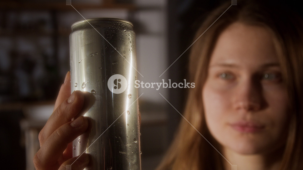 Close up shot of young woman holding drink in aluminum can with no label and pressing cold metal surface to skin refreshing in summer heat indoors and enjoying sensation, copy space