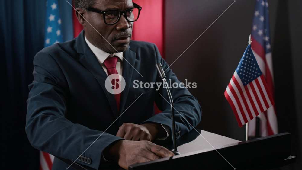 Black man as politician giving passionate speech at podium with American flags in background. Clip is ideal for political campaigns, news broadcasts, and government communications
