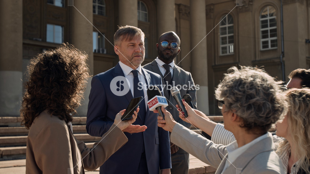 Medium shot with lens flare of mature Caucasian male attorney speaking to journalists with microphones during briefing on court hall steps, protected by security, answering questions on client defense