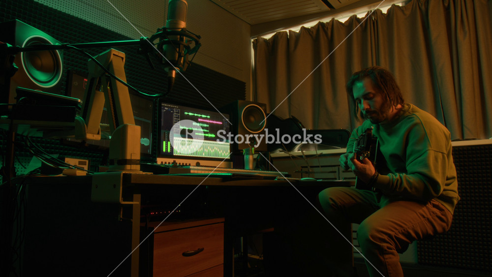 Side full shot of male Caucasian musician creating new song on guitar in green neon lit studio, recording sound into computer with audio sequences on screen for further arrangement