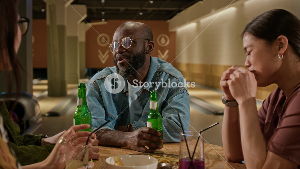 Diverse young adult friends, including African American man, clinking drinks at bowling alley table. Ideal for marketing campaigns promoting social entertainment, nightlife, and friendship