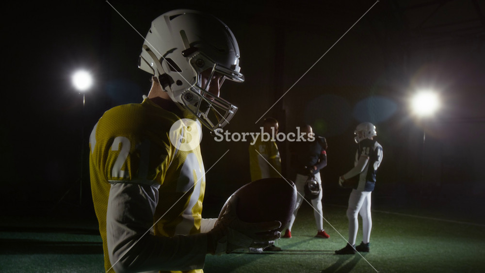 Medium side shot of young Caucasian male gridiron player in yellow uniform with shoulder pads standing in sports arena, bending head and touching helmet with ball before game for prayer or ritual