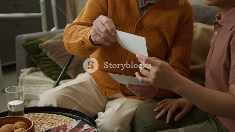 Side view shot of smiling young woman looking at family photos with emotional senior man while sitting on couch at home together and talking, copy space