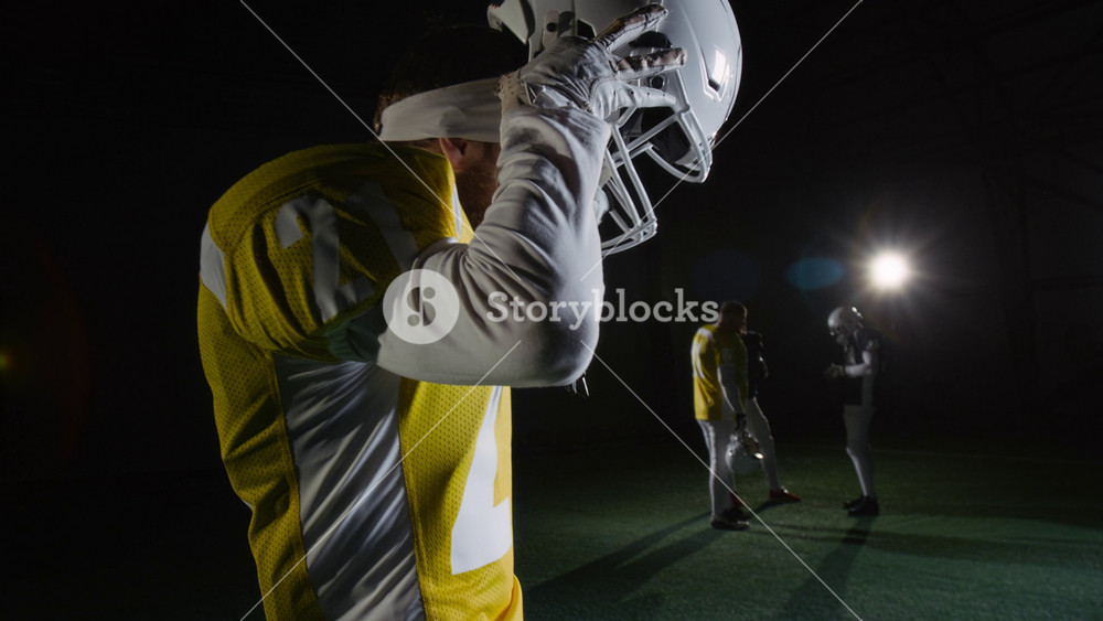 Medium side shot of young male American football player standing in dark arena with spotlights before game practice with team, putting on white protective helmet with mouthguard