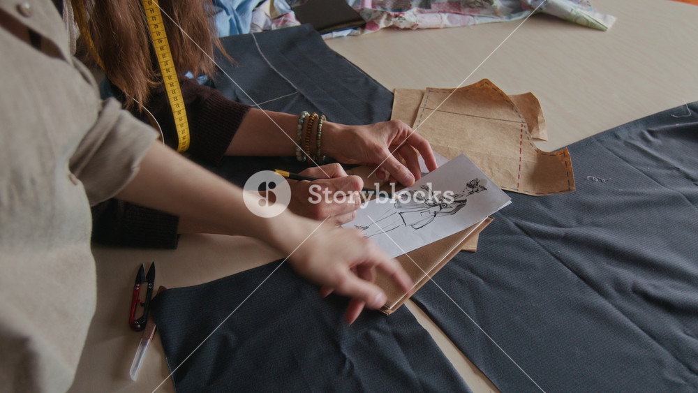 Medium close-up of hands of two unrecognizable female designers drawing fashion sketch of trendy dress model in pencil, suggesting features, while collaborating on new collection in atelier