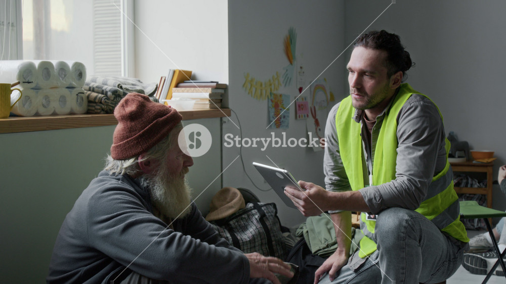 Medium shot of sympathetic young Caucasian male volunteer in yellow vest crouching on floor next to senior refugee man with beard, listening to complaints, then comforting him with hand on shoulder