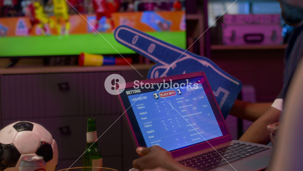 Close up of shot of multiethnic group of friends watching sports match on TV together with focus on hand holding laptop with betting app on screen, copy space