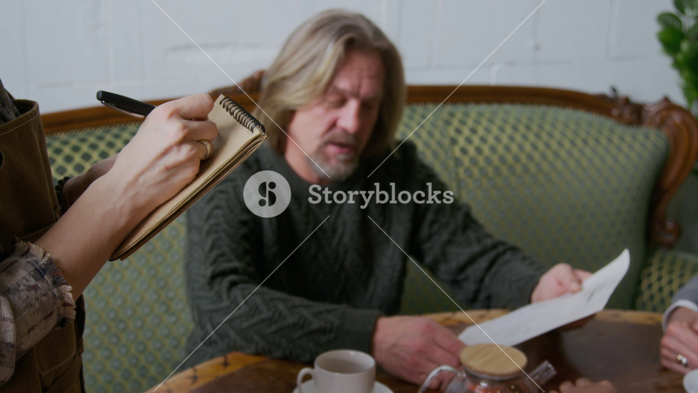 Medium close-up of hands of unrecognizable female waitress in apron writing in notebook while taking lunch order from mature Caucasian male customer, relaxing in company of friends in cafe