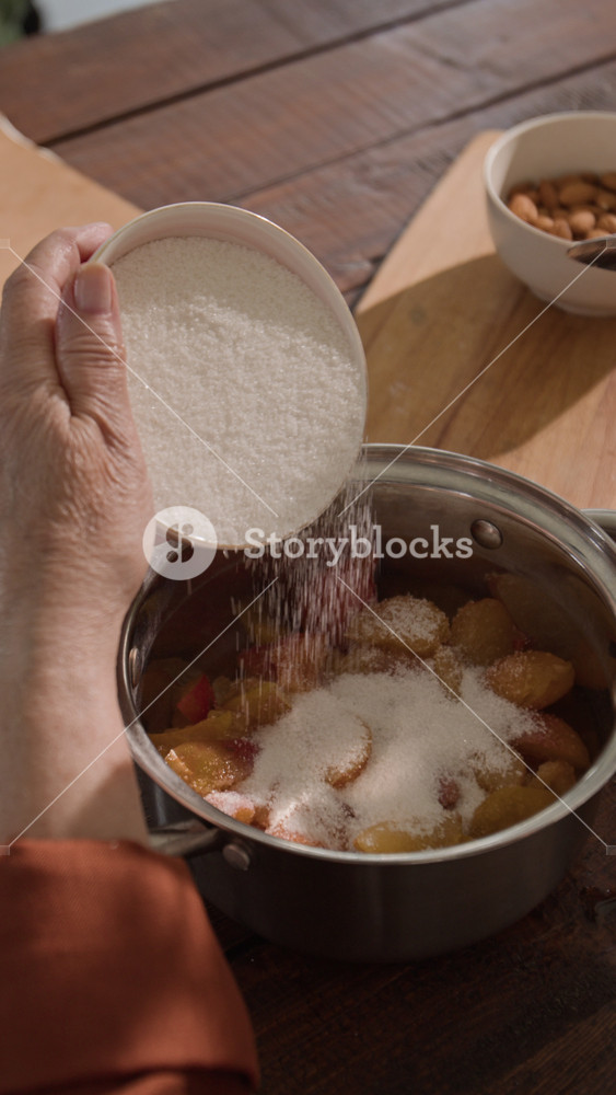 Vertical video of unrecognizable senior woman pouring sugar from bowl into pot with sliced peaches and stirring mixture. Footage is perfect for culinary blogs, recipe ads, and homemade food marketing