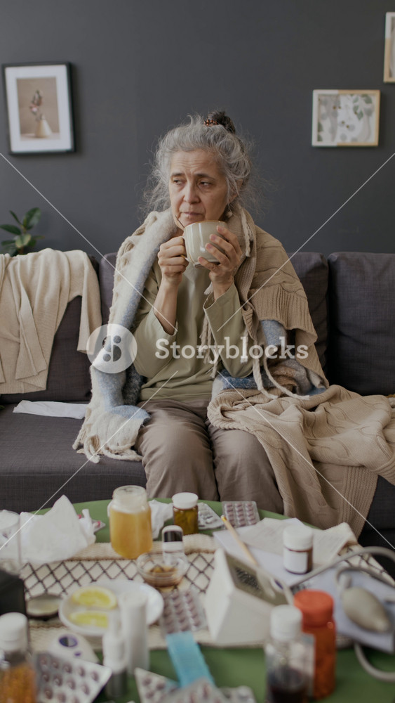 Full vertical shot of senior Caucasian woman with blankets on shoulders drinking cup of hot lemon and honey tea to relieve sore throat and fever while suffering from flu or covid virus