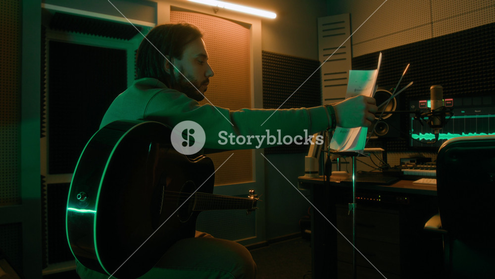 Side shot of young adult Caucasian musician sitting with guitar and putting sheet music on music stand, learning new song or melody in neon lit recording studio