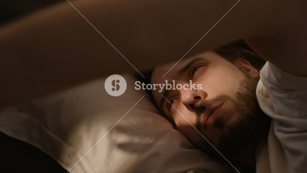 Young Caucasian man waking up in bed, opening bleary eyes and stretching arms in morning. feeling tired. Footage represents sleep health, insomnia, wellness, or remote work lifestyle content
