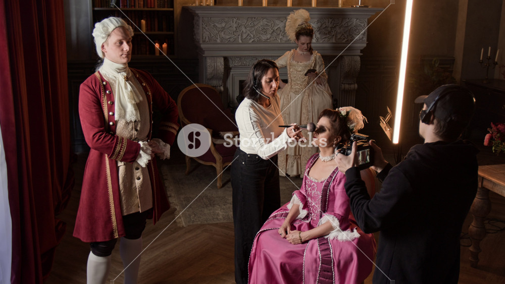 Full shot of film crew and actors working backstage on historical movie set, in aristocratic mansion. Make-up artist powdering face of actress in royal dress, cameraman doing test shots