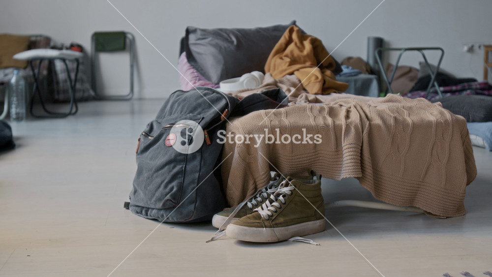 No people medium closeup of backpack, pair of sneakers on floor, next to folding bed with pillow and blankets, in refugee shelter for homeless people displaced due to war or natural disaster