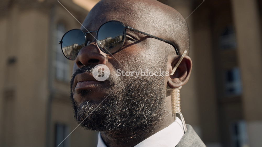 Close-up of face of young African American male security agent or guard, wearing suit and sunglasses, standing outdoors by government office, adjusting transparent earpiece while on duty