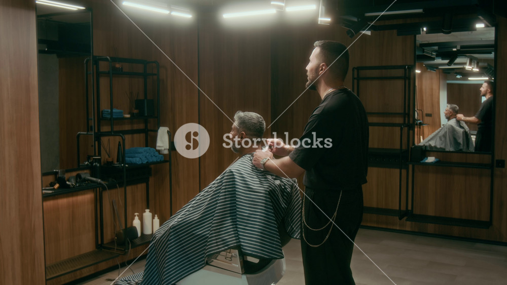 Full shot of young barber styling clients hair after cutting, removing cape, mature Asian man getting up from chair, shaking hands with hairdresser and thanking for good work