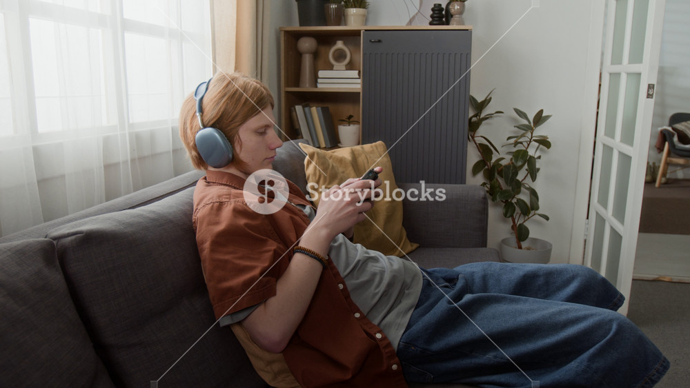 Caucasian teenager wearing headphones sitting on sofa, playing mobile game on smartphone. Clip is suitable for mobile gaming apps, digital entertainment, or telecommunication services marketing