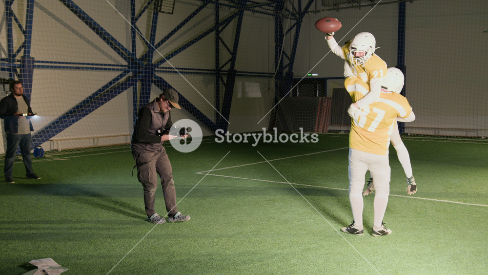 Wide panning shot of cameraman filming gridiron football players in yellow jerseys cheering and hugging after scoring goal during game, while producing content for TV reality show or docuseries