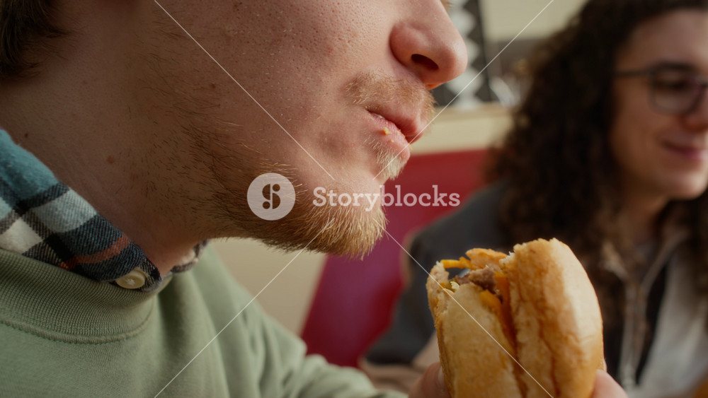 Extreme close-up of lower face of young Caucasian zoomer guy biting off mouthfuls of delicious cheeseburger, chewing with delight and pleasure while indulging in fast food at diner