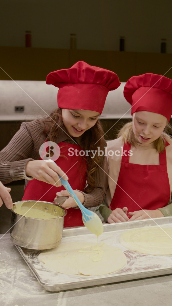 Vertical video of Caucasian children wearing chef hats spreading cream on dough with male instructor in kitchen. Footage is ideal for promoting kids culinary schools or family cooking classes