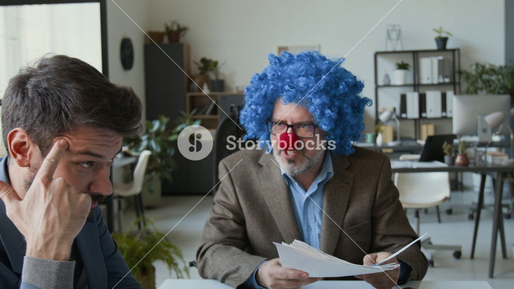 Zoom out shot of clown boss with wig and nose emotionally scolding employees and throwing papers on table, man twisting his finger at temple and looking at female colleague during office meeting