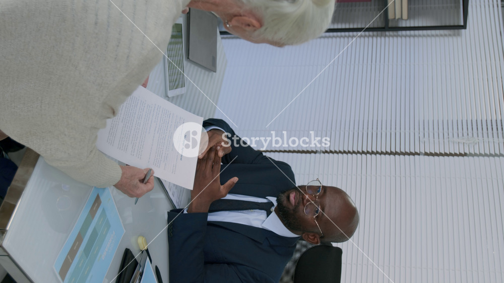 Vertical shot of Black financial advisor sitting at office desk, consulting elderly Caucasian woman on terms of mortgage agreement