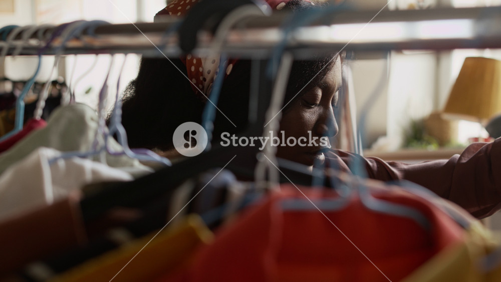 Selective focus close-up of attentive young African American woman browsing clothes on hangers in womenswear section at second hand outlet, examining dress and blouse, searching for stylish bargains