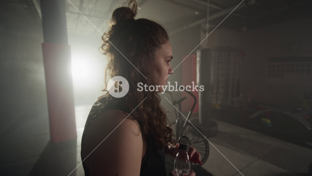 Young Caucasian woman walking into dark gym, removing hoodie, and drinking water from bottle. Footage is ideal for fitness branding, hydration campaigns, or sports advertisements