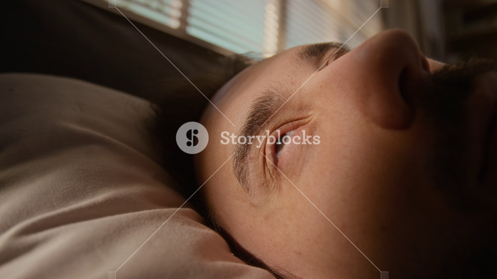Extreme close-up of caucasian young man waking up in bed, opening eyes, looking up, blinking, turning head on pillow. Footage represents morning routine, sleep health, wellness, mental wellbeing