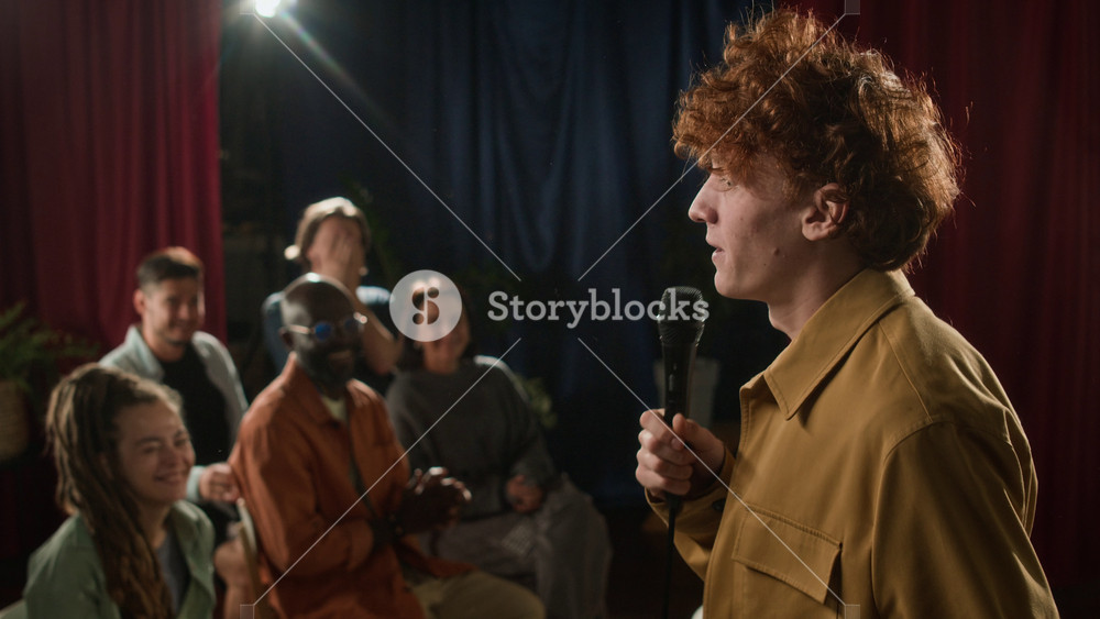 Side low key shot of red-haired male stand-up comedian in yellow shirt gesturing impulsively and sharply while performing on stage and receiving applause from audience