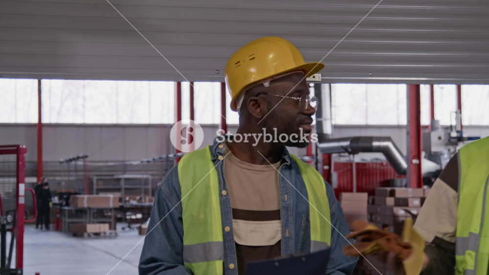 Medium shot of two Asian and African American male colleagues in safety uniforms lifting up roller door, walking out of warehouse, removing hardhats at end of workday, chatting before going home