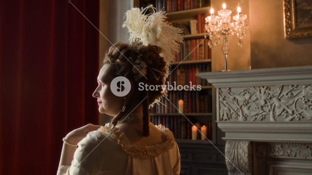 Side view of face of beautiful middle-aged Caucasian noble dame in glamorous historical dress, elaborately styled hair standing by window in castle, looking out while contemplating romantic emotions