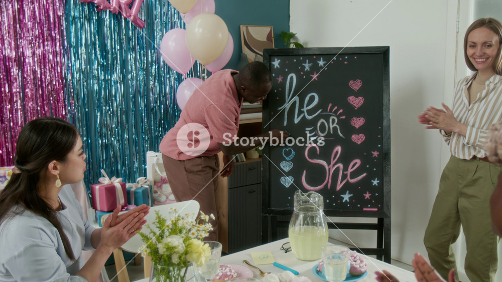 Full zoom-out shot of cheerful African American man drawing hearts and stars on blackboard spelling he or she at gender reveal party, while guests and pregnant wife are watching and clapping
