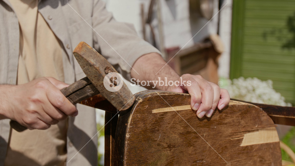 Medium close-up of hands and midsection of thrifty country man restoring old beaten chair in garden, gluing together broken seat, fixing with hammer, displaying practicality and sustainable lifestyle