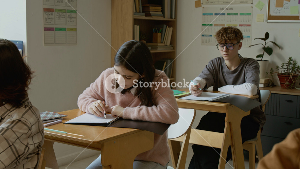 Full shot of motivated Caucasian male and female high school students sitting at desks at lesson, writing test, looking up at blackboard, to achieve good grades for trimester during academic year