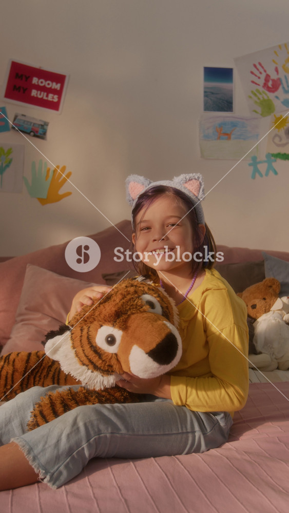 Vertical shot of young girl wearing cat ears headband sitting on pink bed at home and holding big toy tiger smiling at camera